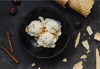 A bowl with three scoops of ice cream, topped with nuts. Surrounding the bowl are waffle scraps and some cinnamon sticks.