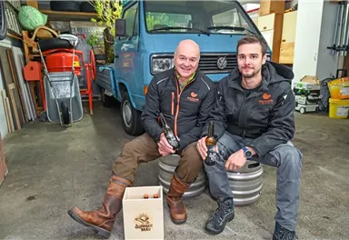 Zwei Männer sitzen in einer Garage auf Fässern und halten Bierflaschen in der Hand. Im Hintergrund steht ein blauer Volkswagen-Bus.