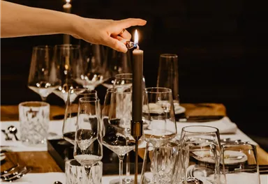 Ein eleganter Esstisch mit Gläsern und Besteck. Eine Hand zündet eine Kerze an und sorgt für gemütliches Licht.