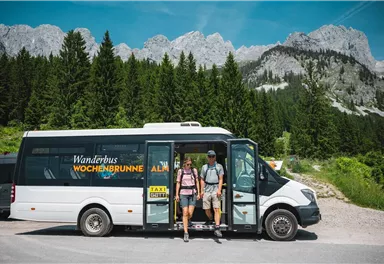 Ein Wanderbus steht in der Natur, umgeben von Bergen und Wäldern. Zwei Personen steigen aus, bereit für ein Abenteuer.