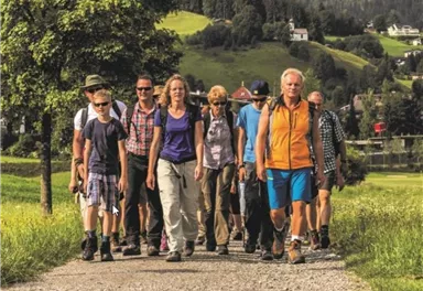 A group of hikers walks along a narrow path, surrounded by green hills. The participants are wearing hiking clothes and seem to be enjoying nature.