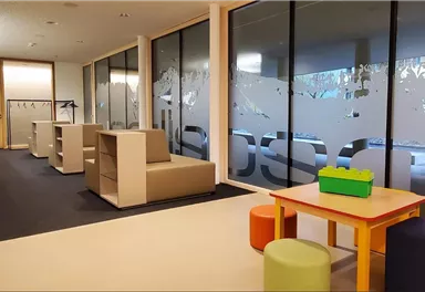 A modern room with seating and large windows. In the center stands a colorful table with chairs for children.