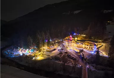 A picturesque winter landscape with festively illuminated buildings and trees. The scene depicts a cozy atmosphere at night.