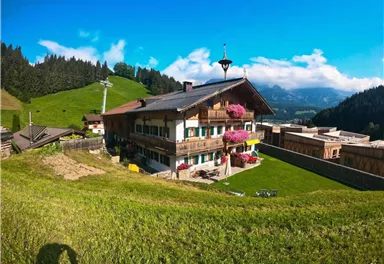 Ein schönes alpenländisches Haus mit bunten Blumenbalkonen. Im Hintergrund erstreckt sich eine grüne Wiese und bewaldete Hügel unter einem blauen Himmel.
