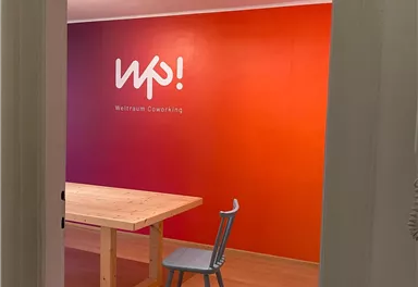 A friendly coworking space with a large table and a chair. The wall is designed in warm colors and features the logo "wp!".