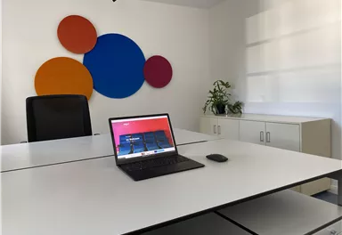 A modern office with a large white table and a laptop. Colorful circles are on the wall, and a plant is visible.