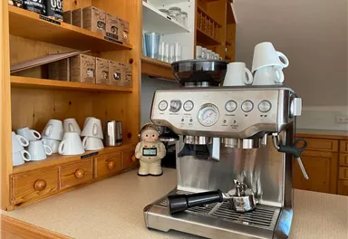 A modern coffee machine is placed on a countertop. In the background, various coffee cups and a decorative figure are visible.