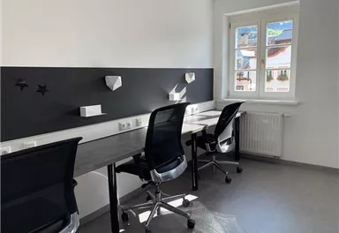 A modern office with bright walls and large windows. There are several desks with chairs and a minimalist design.