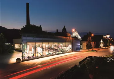 Ein modernes Gebäude mit großen Fenstern, das bei Nacht beleuchtet ist. Im Vordergrund sind Straßenlichter und Pflanzen zu sehen.