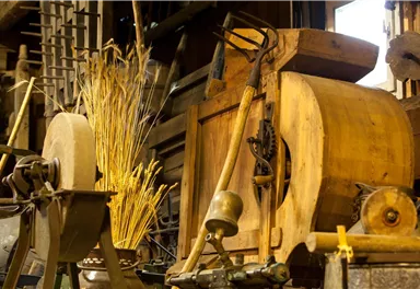 Ein historisches Handwerksgerät mit Holzdetails steht in einem alten Schuppen. Neben dem Gerät sind getrocknete Halme und verschiedene Werkzeuge zu sehen.
