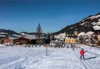 A winter landscape with snow-covered hills and a small town in the background. People are skiing and enjoying the clear blue sky.