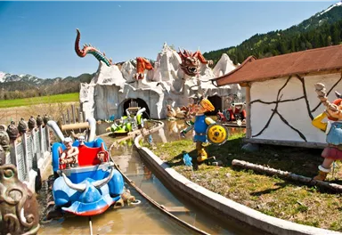 A theme park with a water ride and colorful figures. In the background, mountains and a blue sky can be seen.