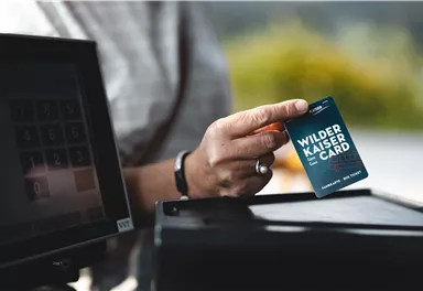 A person is holding a Wilder Kaiser Card in their hand. In the background, a cash register terminal is visible.