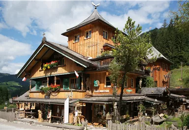Ein traditionelles, holzverkleidetes Alpenhaus mit vielen bunten Blumen und einer malerischen Umgebung. Der Himmel ist blau mit einigen Wolken und grünem Wald im Hintergrund.