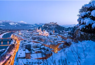 Eine malerische Stadt im Winter, bedeckt mit Schnee und umgeben von Bergen. Die Lichter der Stadt reflektieren sich im ruhigen Wasser des Flusses.