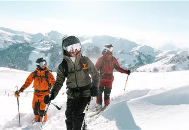 Drei Menschen wandern durch eine verschneite Berglandschaft. Die Sonne scheint, und die Umgebung ist malerisch mit hohen Bergen im Hintergrund.