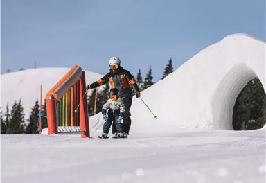 Ein Skilehrer hilft einem Kind beim Skifahren auf einer schneebedeckten Piste. Die Szene zeigt ein sonniges Wetter mit einer verschneiten Landschaft im Hintergrund.