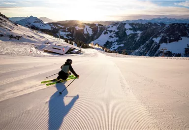 Ein Skifahrer fährt eine breite, sonnige Piste hinunter. Im Hintergrund sind schneebedeckte Berge und ein klarer Himmel zu sehen.