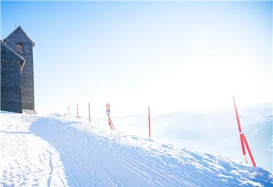 Eine schneebedeckte Landschaft mit einer kleinen Kirche und roten Markierungen. Der Himmel ist hell und klar, während die Berge im Hintergrund verschwommen sind.