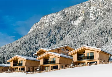 A modern wooden house in the snow against an impressive mountain backdrop. The sky is blue and the landscape is peaceful and wintry.