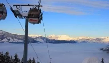 A cable car glides over a forested mountainous landscape. In the background, snow-covered mountains and a sea of fog can be seen.