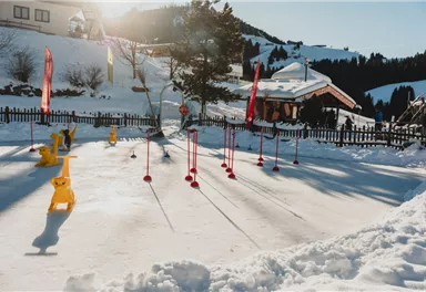 Ein fröhliches Kind in einem roten Schneeanzug fährt auf Skiern. Es steht auf einer verschneiten Piste mit einem blauen Himmel im Hintergrund.