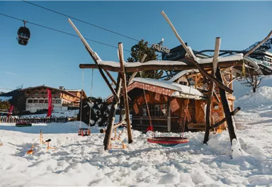 Zwei Personen in Winterkleidung stehen auf einer Eisbahn. Sie halten Besen und sitzen auf bunten Schlittenfiguren.