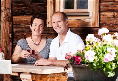 Ein Paar sitzt an einem Holztisch vor einer rustikalen Hütte. Sie lächeln und halten ein Glas in der Hand, umgeben von bunten Blumen.