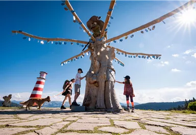 Eine Gruppe von Menschen steht um eine große Statue mit vielen Armen. Im Hintergrund sind Berge und ein Leuchtturm zu sehen, und die Sonne scheint.