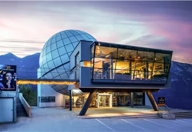 A modern building with a glass facade and a spherical structure at the top. In the background, there are gentle hills and a colorful sky.