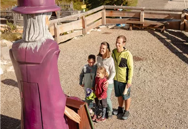 Eine Gruppe von Kindern steht vor einer bunten Zaubererfigur im Freien. Im Hintergrund sind grüne Wiesen und einige Gebäude zu sehen.