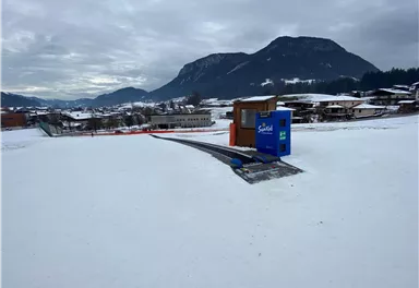 A snowy landscape with a small wooden house and a blue box. In the background, mountains and some houses can be seen.