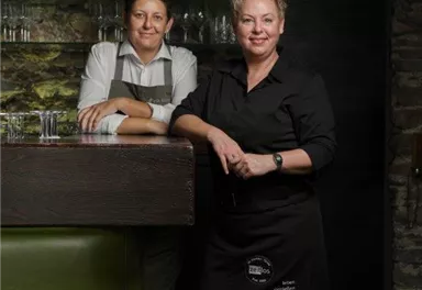 Zwei Personen stehen an einer Bar in einem gemütlichen Restaurant. Im Hintergrund sind Regale mit Getränken zu sehen.