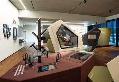 A modern exhibition room with interactive displays and exhibits. There are several monitors, headphones, and geometric structures on the table.