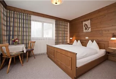 Ein gemütliches Schlafzimmer mit einem Doppelbett und Holzverkleidung. Neben dem Bett steht ein kleiner Tisch und Stühle, und ein Fenster mit Vorhängen sorgt für natürliches Licht.