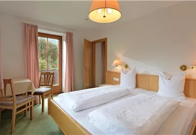 Ein helles Zimmer mit zwei Einzelbetten und einer gemütlichen Einrichtung. Der Raum verfügt über ein Fenster mit Blick nach draußen und einen kleinen Tisch mit Stühlen.