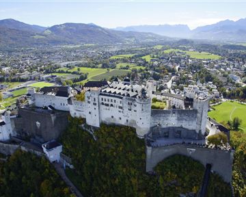 An impressive castle on a hill, surrounded by green meadows and mountains. The clear sky and picturesque landscape create an idyllic atmosphere.