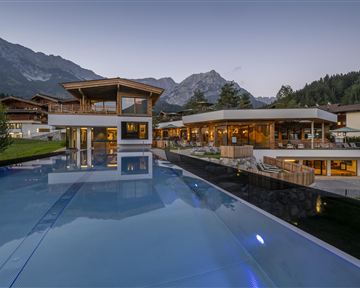 A modern building with large windows and a beautiful infinity pool. In the background, majestic mountains and trees are visible.