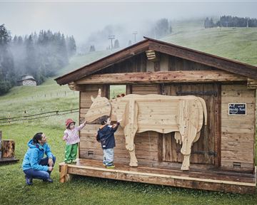 Ein Holzhaus mit einer Holzfigur einer Kuh steht auf einer Wiese. Zwei Kinder und eine Frau stehen davor und erkunden die Figur.