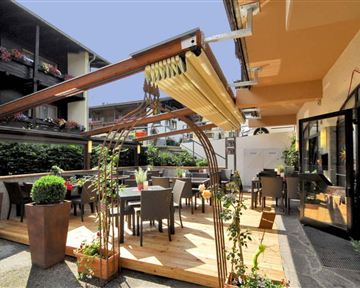 Ein gemütlicher Außenbereich eines Restaurants mit Holzterrasse und Tischen. Umgeben von Pflanzen und einem klaren Himmel.
