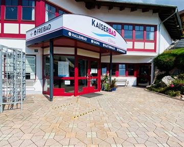 A building with the inscription "Kaiserbad" and a large entrance door.  
In front of the entrance are paved paths and some plantings.