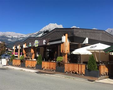Ein gemütliches Restaurant mit einer Holzterrasse und Sonnenschirmen. Im Hintergrund sind majestätische Berge und ein blauer Himmel zu sehen.