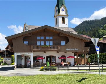 A traditional alpine inn with a characteristic clock tower. In the foreground, there are tables with red umbrellas and a green meadow.
