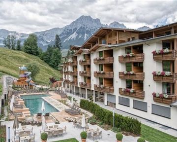 A modern hotel with wooden cladding and balcony flowers. In the background, majestic mountains and a swimming pool with loungers can be seen.