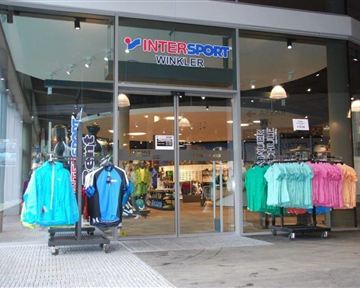 A store entrance with the name "INTER SPORT WINKLER". In front of the door, there are several clothing racks with colorful sportswear.
