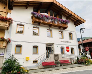 A beautiful building with a balcony adorned with flowers. It is the municipal office with an inviting staircase and two red benches in front.