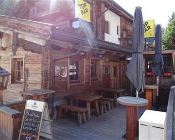 A rustic cabin with wooden cladding and an outdoor area. There are tables, umbrellas, and a sign that reads "Top Bar."