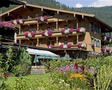 Ein hübsches Holzhaus mit Balkonen, die mit bunten Blumen geschmückt sind. Im Vordergrund gibt es einen schönen Garten mit vielen Pflanzen und Blumen.