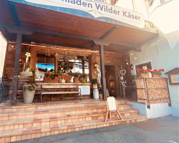 Ein einladender Bauernladen mit dem Schild "Wilder Käser" über dem Eingang. Der Eingangsbereich ist mit Pflanzen dekoriert und hat eine gemütliche Sitzgelegenheit.