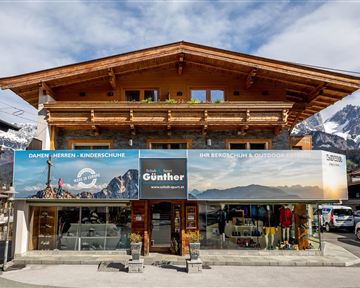 Ein altes Holzgebäude, das ein Geschäft für Outdoor-Ausrüstung beherbergt. Der Laden zeigt großflächige Werbebanner und befindet sich in einer bergigen Umgebung.